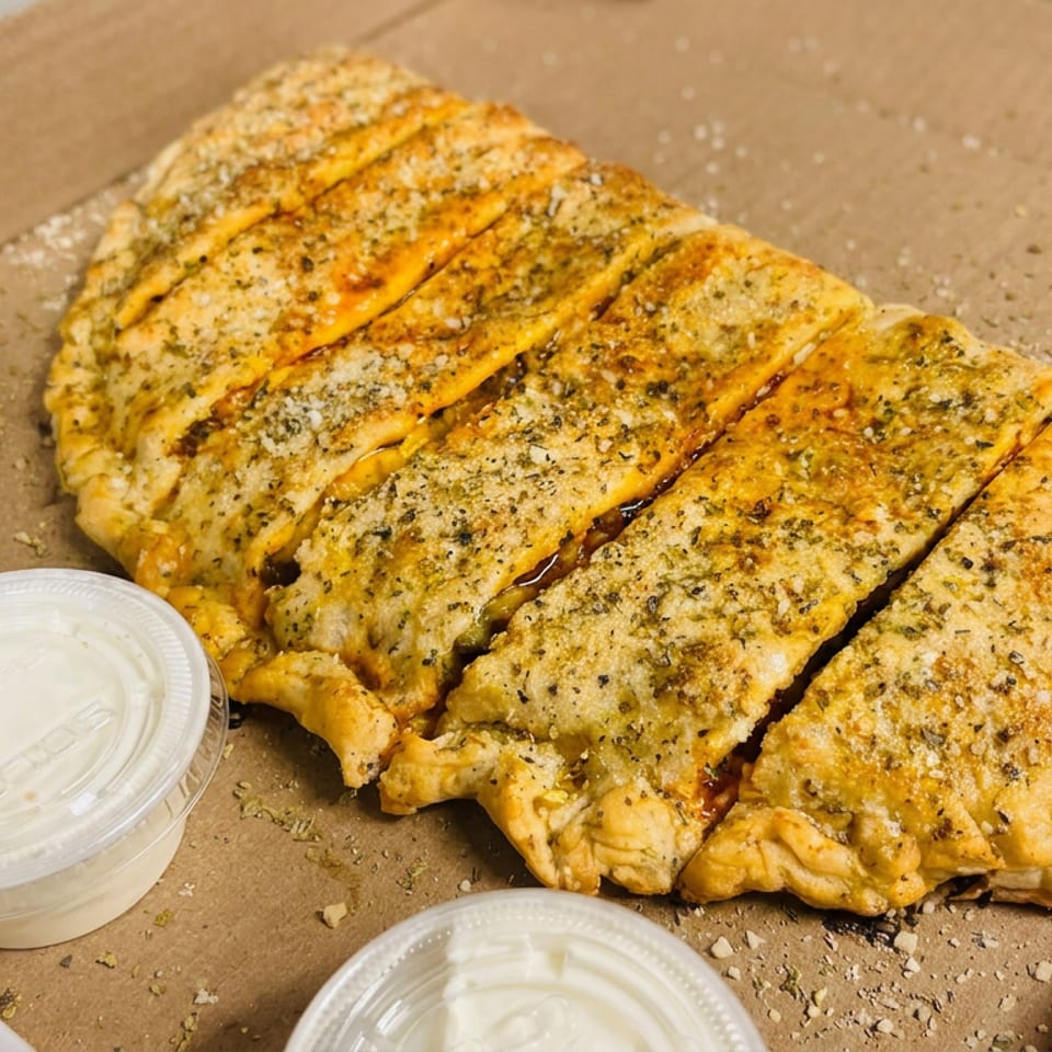 Sliced golden garlic bread topped with herbs and cheese, served in a cardboard box with three cups of creamy dipping sauce.